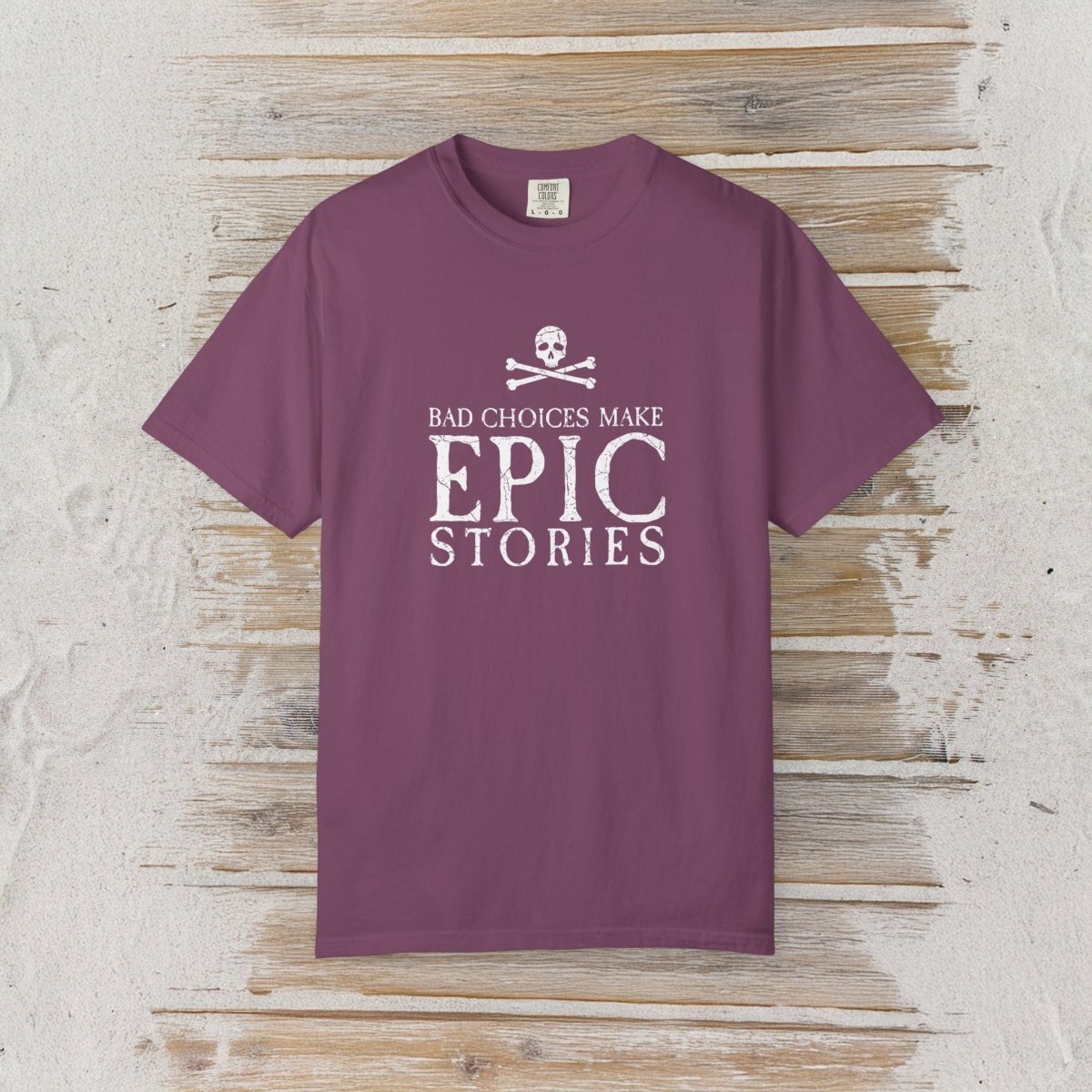 A short-sleeve crewneck t-shirt in a dusty plum purple color, featuring a white graphic print on the front with a skull and crossbones and the text 'BAD CHOICES MAKE EPIC STORIES'. The shirt is displayed flat against a rustic, whitewashed wooden plank background.