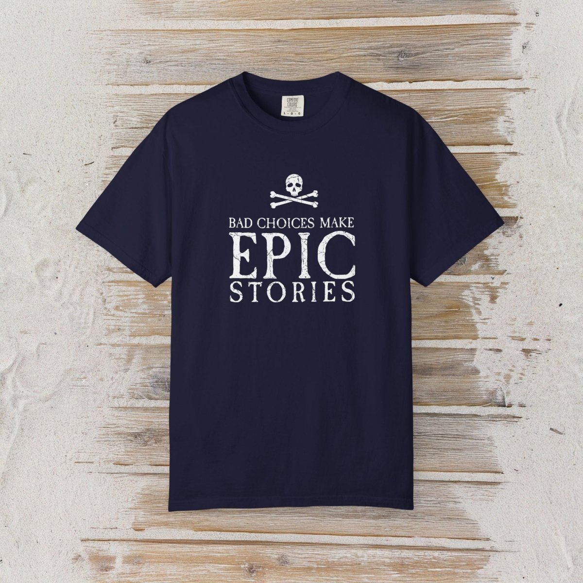 A navy blue short-sleeve t-shirt with a white graphic print on the front, laid flat on a rustic light wood plank background. The graphic features a skull and crossbones above the text 'BAD CHOICES MAKE EPIC STORIES'.