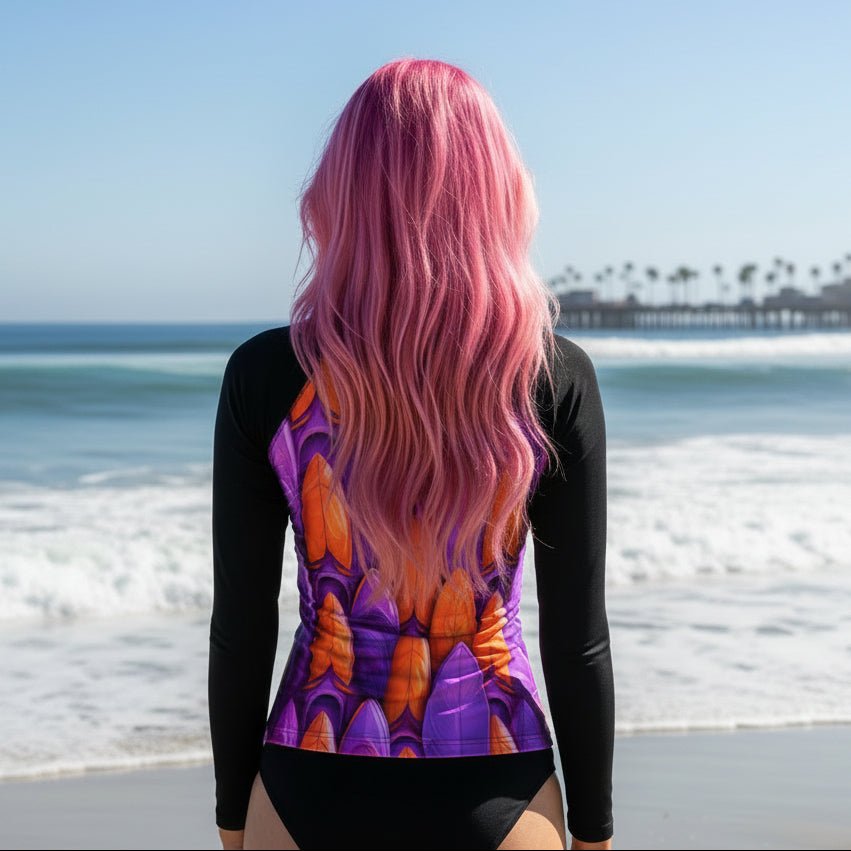 A woman with long pink hair stands on a beach facing the ocean, and the pismo beach pier, wearing a long-sleeve swimsuit with black sleeves and a purple and orange abstract surfboard patterned back panel.