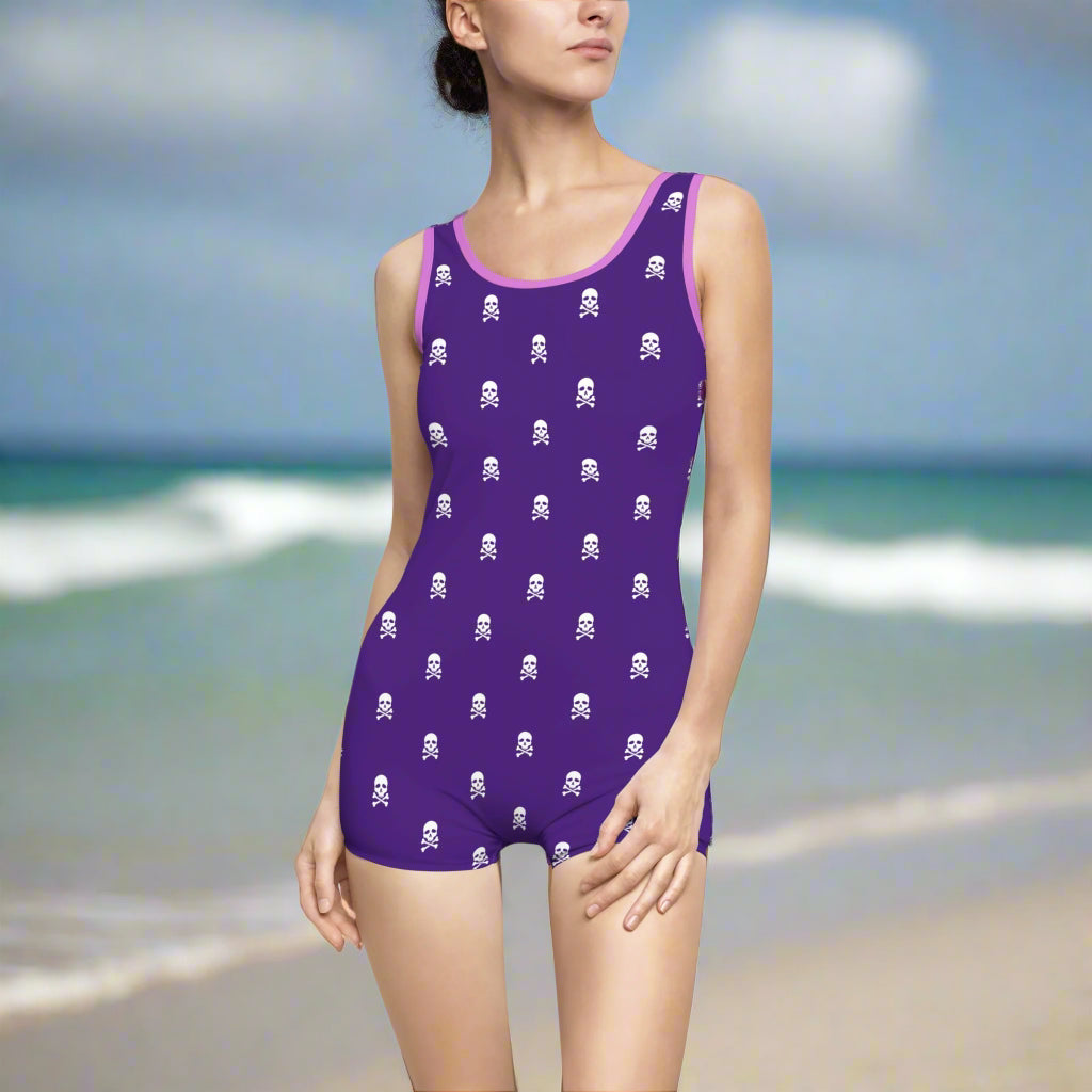 Model wearing a fun purple one piece bathing suit with pirate skull polka dots on the beach