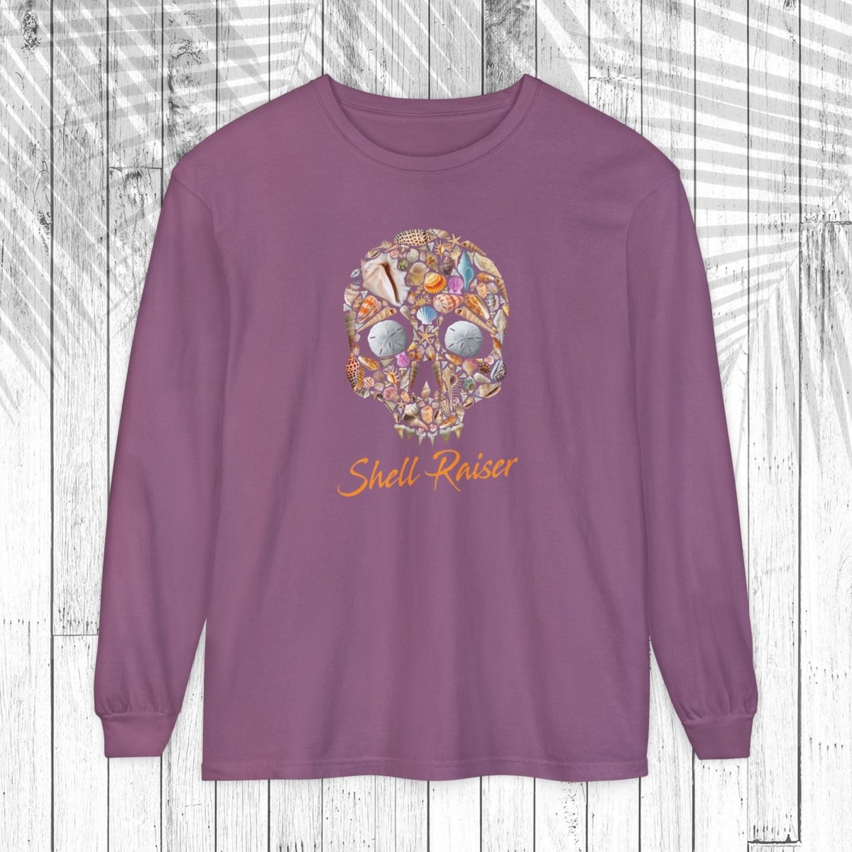 A long-sleeve crewneck t-shirt in dusty purple, featuring a colorful graphic of a skull made of various shell illustrations and the words 'Shell-Raiser' printed below in orange script, displayed on a white wood plank background.
