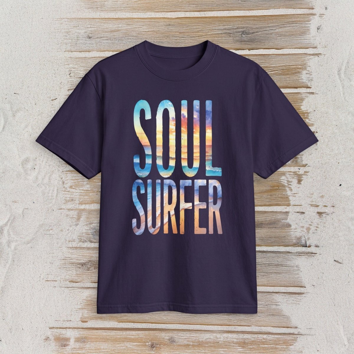 A dark purple short-sleeve t-shirt with 'SOUL SURFER' printed in a colorful gradient block-letter font, displayed flat on a light wood plank background.