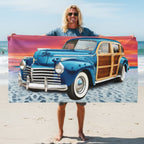 Surfer dude holding up a rectangular beach towel featuring a Vintage Woody Wagon on a sandy beach under a colorful sunset sky.