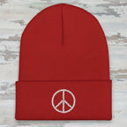 A red ribbed knit beanie hat with a folded cuff, featuring a white embroidered peace sign on the front, shown against a plain white background.