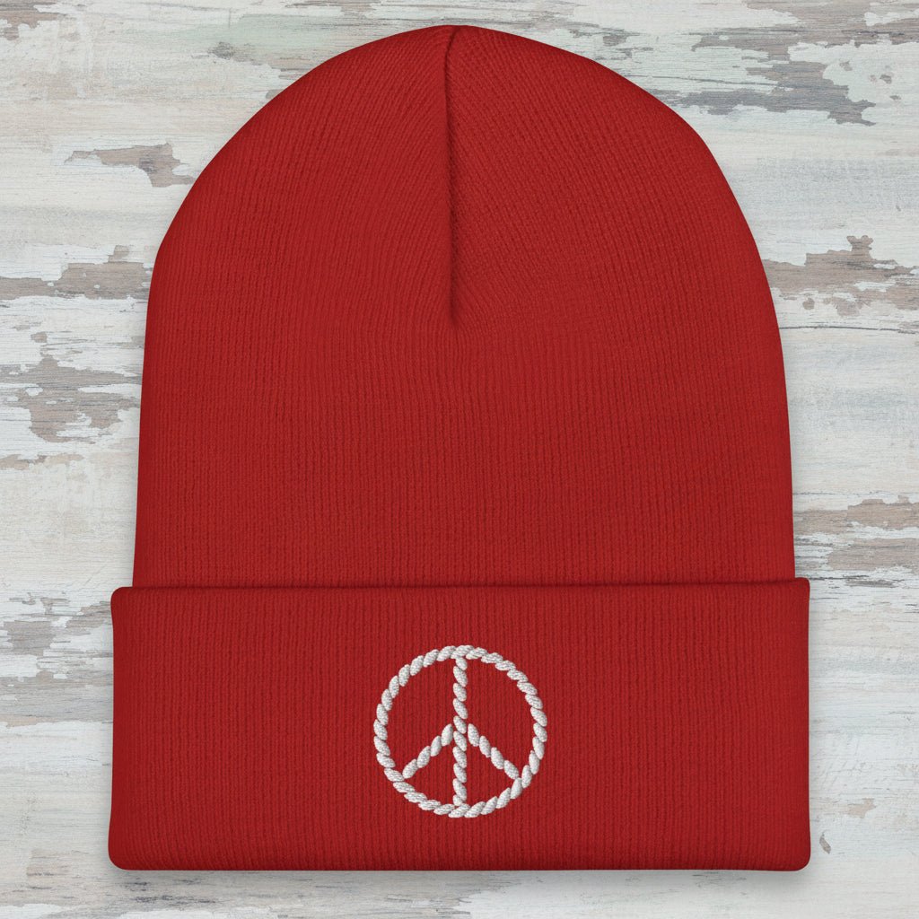 A red ribbed knit beanie hat with a folded cuff, featuring a white embroidered peace sign on the front, shown against a plain white background.