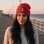 A red knit beanie hat with a folded cuff featuring an embroidered gray and white skull and crossbones design, shown against a plain white background.