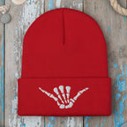 A red ribbed knit beanie hat with a folded cuff featuring a white embroidered shaka hand symbol, shown on a rustic wood background