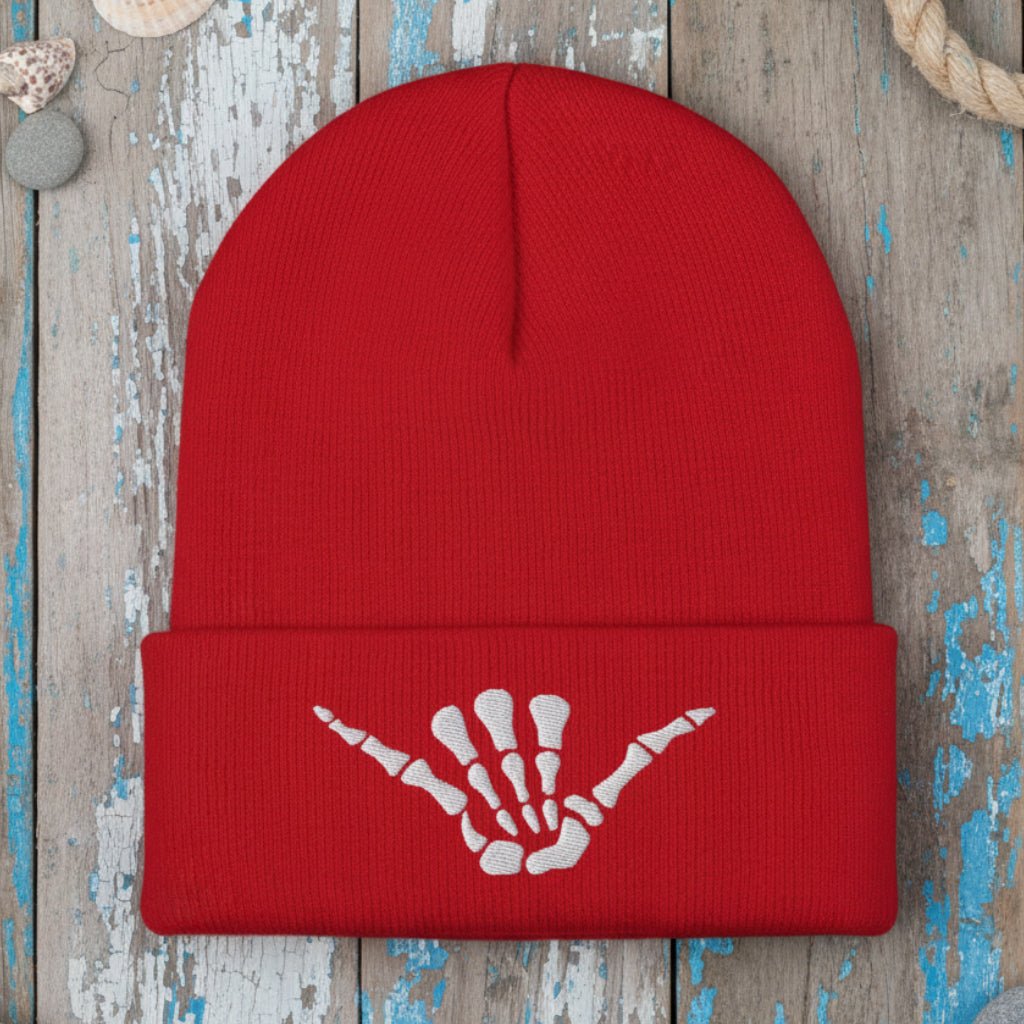 A red ribbed knit beanie hat with a folded cuff featuring a white embroidered shaka hand symbol, shown on a rustic wood background