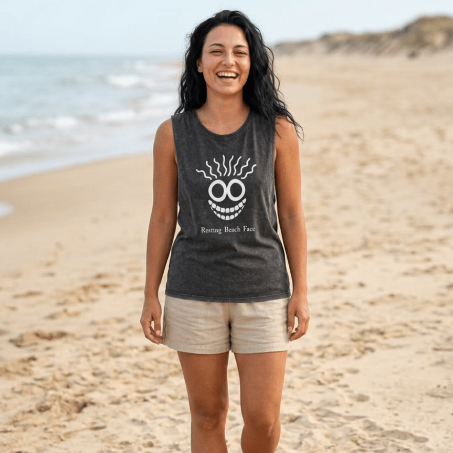 A woman walking on a sandy beach wearing a dark charcoal gray sleeveless tank top with a white graphic of a smiling face with smoke-like hair and the text 'Racing Back Face', paired with light beige shorts.