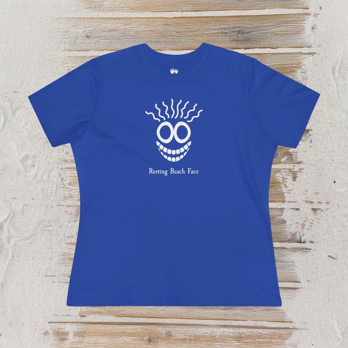 A royal blue short-sleeve t-shirt laid flat on a light wood background. The shirt features a white graphic of a smiling face with wavy hair and the text 'Resting Beach Face' printed below it.