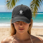 A woman on a beach wearing a dark gray baseball cap with a small white sea turtle embroidered on the front, with turquoise ocean and palm trees in the background.