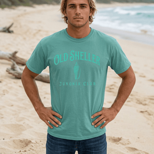 A man standing on a Sanibel Island beach wearing a teal t-shirt with 'OLD SHELLER JUNIORIA CLUB' printed in teal on the front, paired with dark blue jeans.