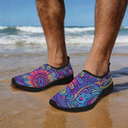 A person standing on a sandy beach wearing slip-on water shoes with a vivid psychedelic floral and paisley pattern in purple, blue, pink, orange, and yellow. The ocean waves and blue sky are blurred in the background.