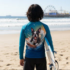 A person standing on Santa Monica Beach California facing the ocean, wearing a blue long-sleeve rash guard with a large graphic print of a Boston Terrier surfing on the back, and holding a white surfboard.
