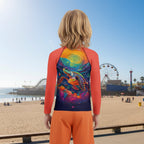 A child stands on a beach boardwalk with their back to the camera, wearing a long-sleeve swim shirt with bright coral sleeves and a colorful ocean turtle graphic on the back, paired with orange swim trunks. The background shows a sunny beach, ocean, and Santa Monica Pier.