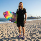 Surfer dude with long stringy blond hair wearing water shoes and black board shorts holding a surfboard standing on Santa Monica Beach - in square format 