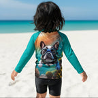 A child standing on a Sarasota Siesta Kee beach facing the ocean, wearing a teal long-sleeve rash guard with a large graphic print of a French bulldog wearing scuba gear on the back, paired with black swim shorts.