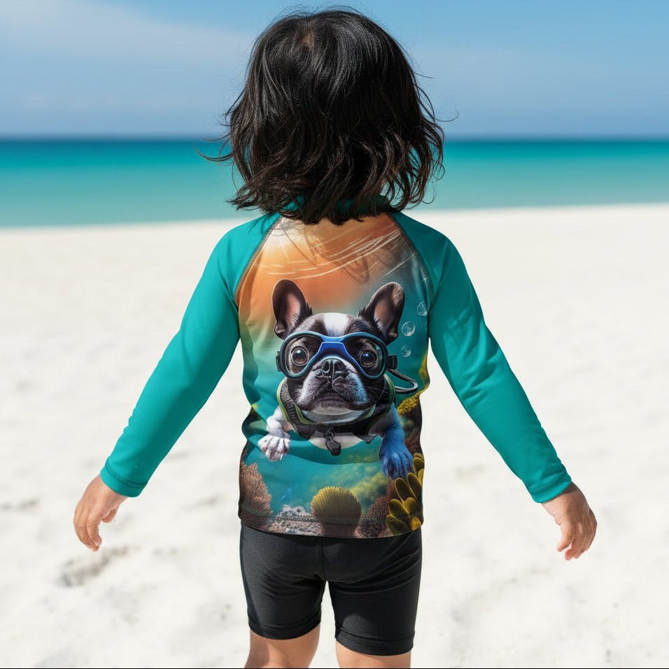 A child standing on a Sarasota Siesta Kee beach facing the ocean, wearing a teal long-sleeve rash guard with a large graphic print of a French bulldog wearing scuba gear on the back, paired with black swim shorts.