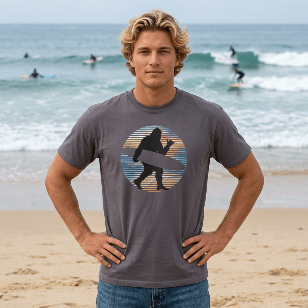 A man standing on a sandy beach wearing a dark gray t-shirt with a circular graphic of a surfing sasquatch silhouette against a striped sunset. The background shows the ocean with surfers and waves.