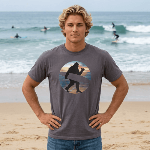 A man standing on a sandy beach wearing a dark gray t-shirt with a circular graphic of a surfing sasquatch silhouette against a striped sunset. The background shows the ocean with surfers and waves.