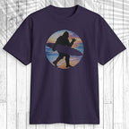 A dark navy short-sleeve t-shirt with a retro circular graphic of a surfer silhouette, displayed flat against a white wood plank background.