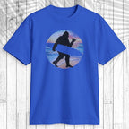 A royal blue short-sleeve t-shirt with a graphic of a black silhouette of Bigfoot carrying  surfboard inside a circular retro sunset design with blue, orange, and red stripes, displayed on a white wood plank background.