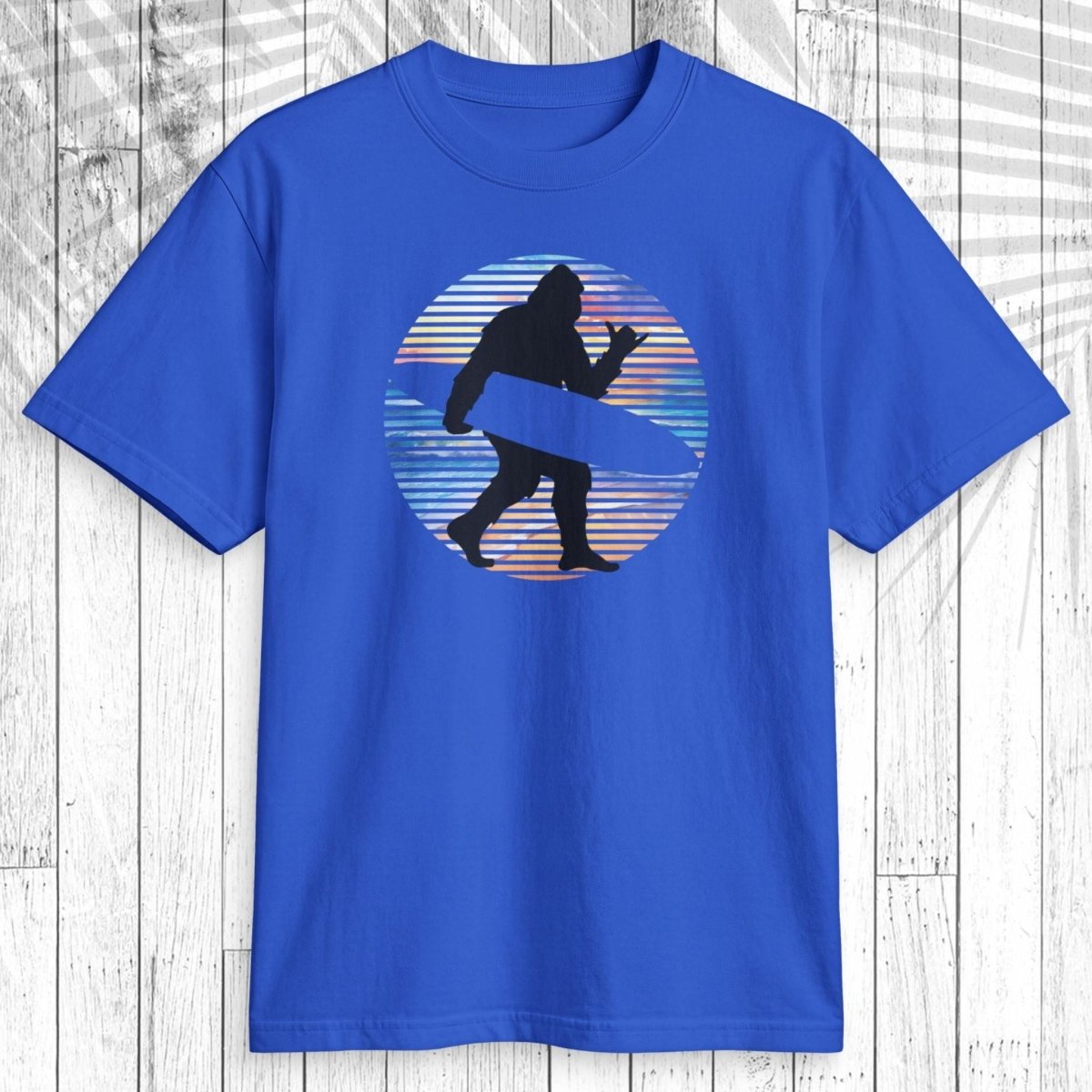 A royal blue short-sleeve t-shirt with a graphic of a black silhouette of Bigfoot carrying  surfboard inside a circular retro sunset design with blue, orange, and red stripes, displayed on a white wood plank background.