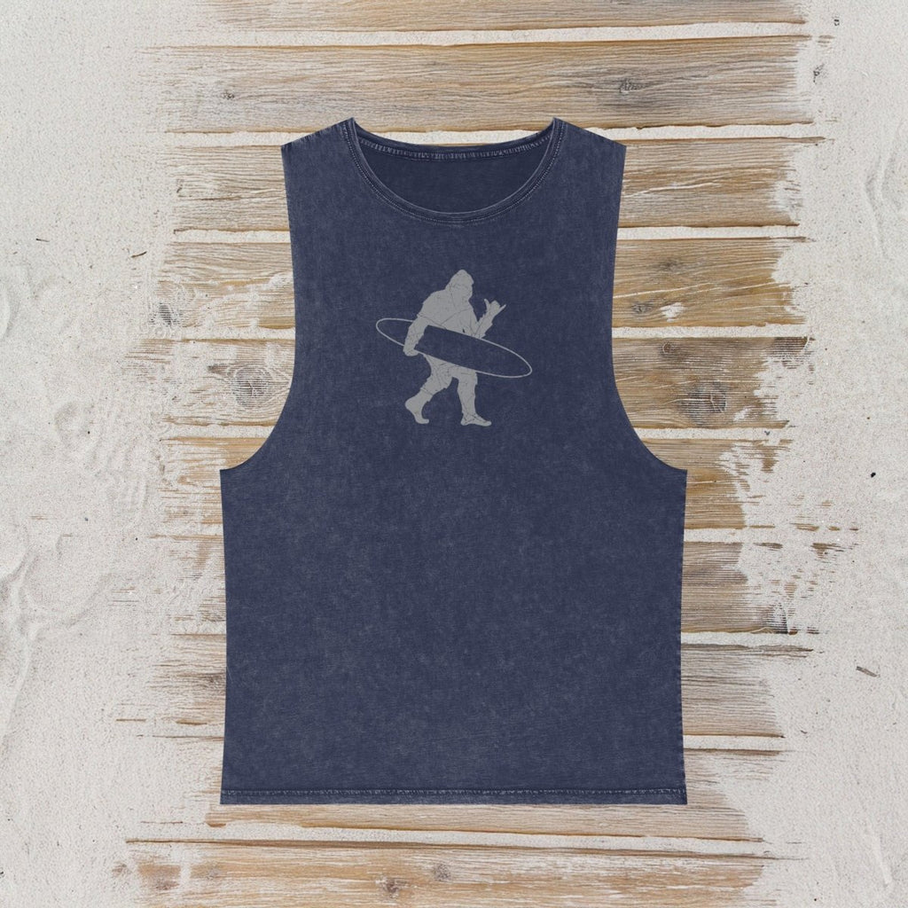 A sleeveless tank top in a washed navy blue color, featuring a light gray graphic of a Bigfoot silhouette carrying a surfboard, laid flat on a rustic wooden plank background.