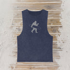 A sleeveless tank top in a washed navy blue color, featuring a light gray graphic of a Bigfoot silhouette carrying a surfboard, laid flat on a rustic wooden plank background.