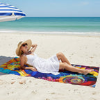 A woman in a white dress and straw hat relaxes on a colorful seahorse beach towel on a sandy beach under a blue and white striped umbrella.