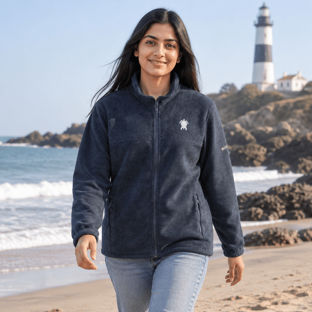 A woman walking on a sandy beach wearing a dark navy fleece zip-up jacket with a small white turtle logo on the left chest, paired with light blue jeans. A lighthouse is visible in the background.