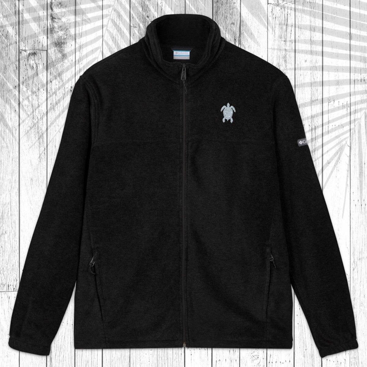 A dark gray fleece zip-up jacket with a stand collar, shown flat against a white wood plank background with a palm leaf pattern overlay. The jacket features a small white circular logo on the left chest and a small white logo on the right sleeve.