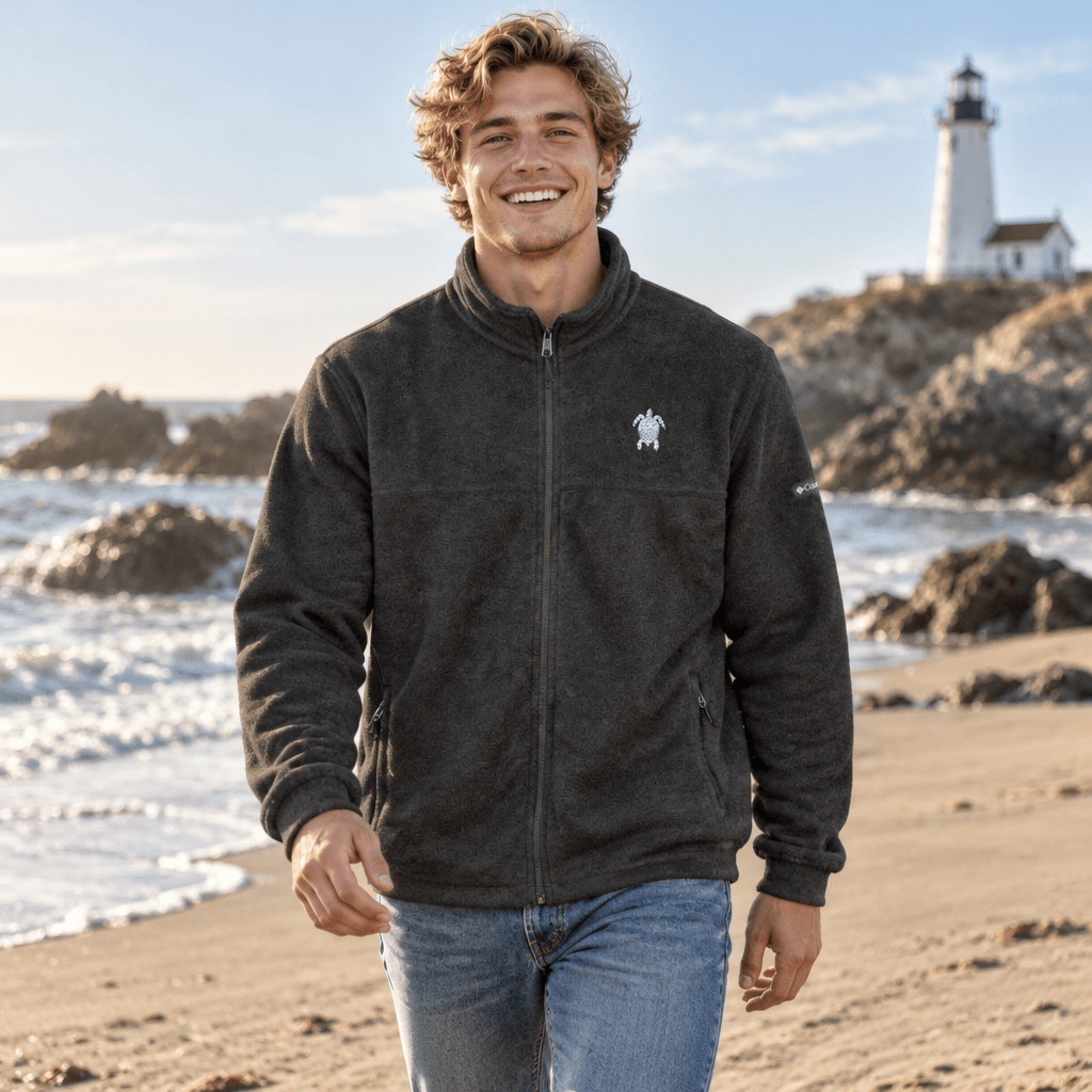A smiling man wearing a dark charcoal fleece zip-up jacket with a small white turtle logo on the left chest, standing on a sandy beach with ocean waves and a lighthouse in the background.