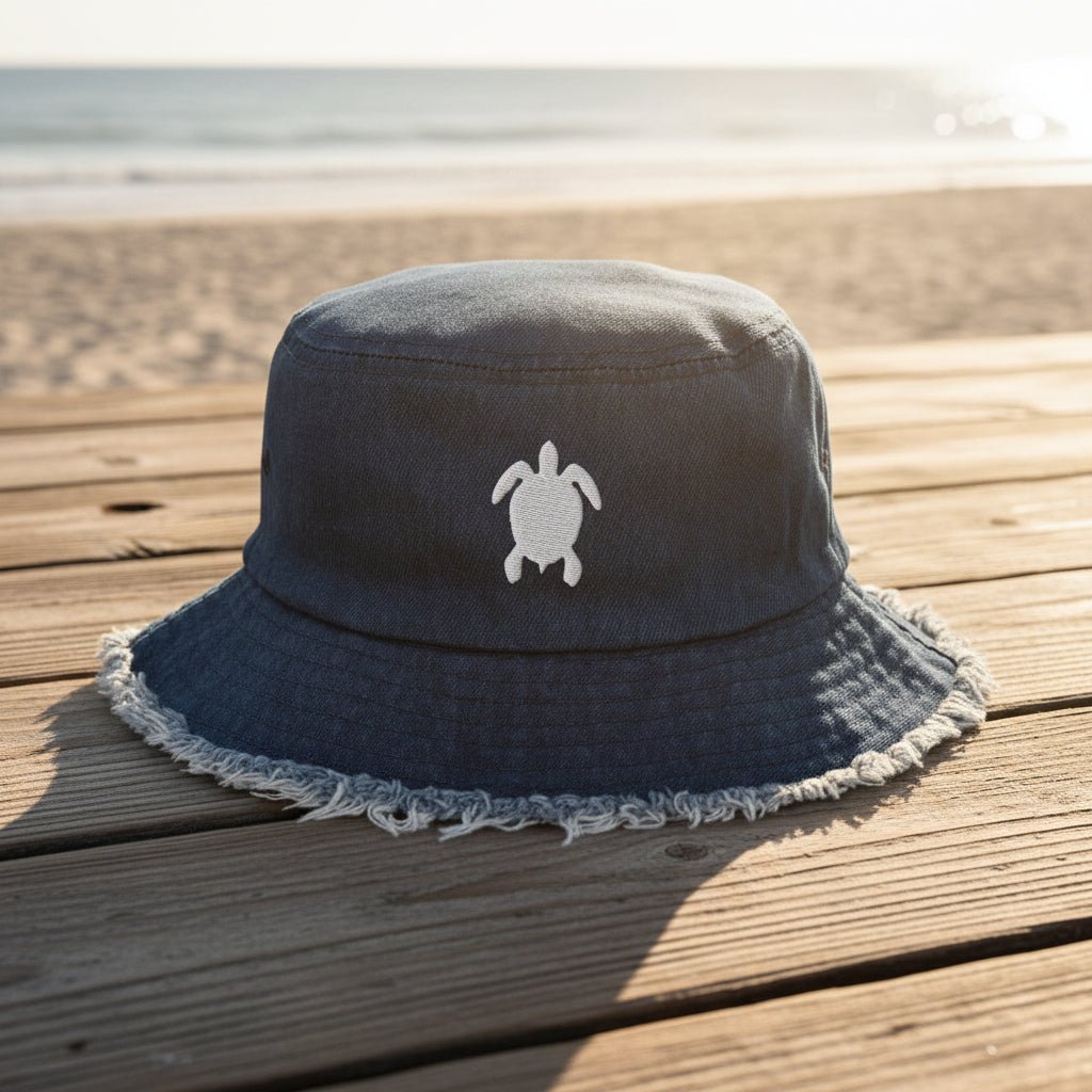 Navy blue bucket hat with white fringe on and a sea turtle embroidery sitting on a beachside boardwalk