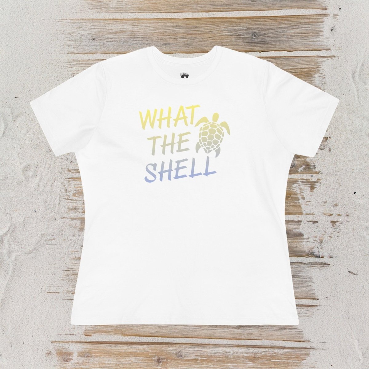 A white short-sleeve t-shirt with a graphic print reading 'WHAT THE SHELL' in yellow and blue gradient text next to a sea turtle illustration, laid flat on a rustic wooden surface.