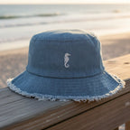Blue denim bucket hat with a seahorse logo on a beachside boardwalk