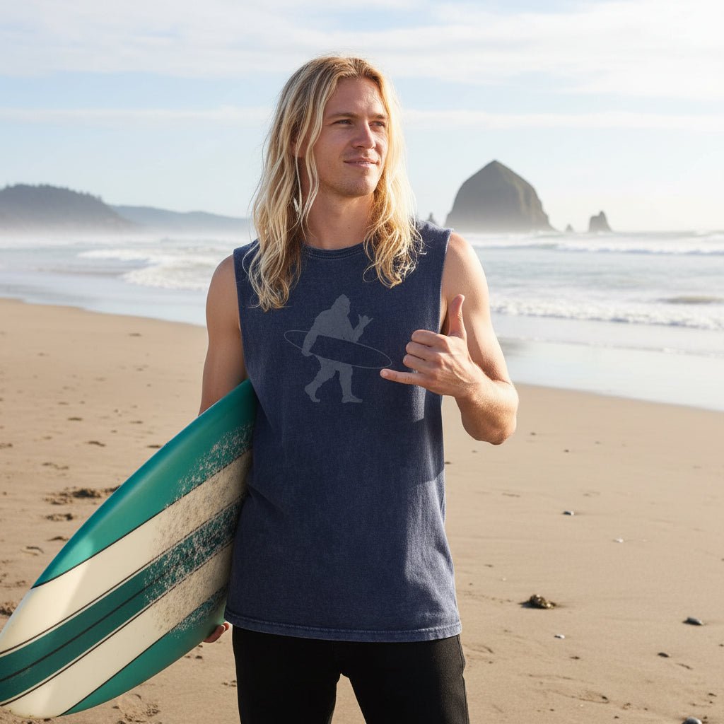 A man wearing a sleeveless navy blue t-shirt with a gray graphic of surfing Sasquatch standing on the beach in seaside oregon