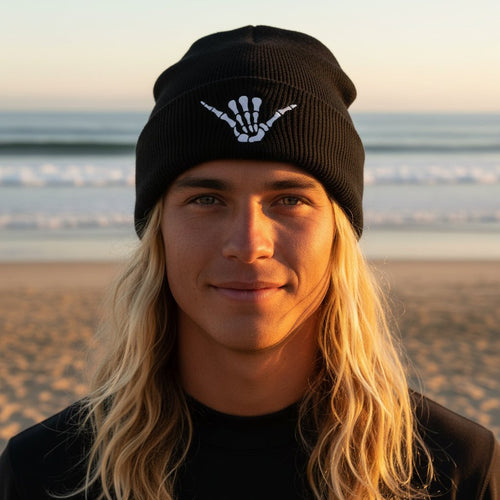 Surfer wearing a black knit beanie hat with a white shaka sign skeleton hand graphic on the folded cuff, standing on La Jolla beach