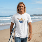 A man walking on a sandy beach holding a surfboard under his arm, wearing a white Hang Loose Shaka t-shirt with a colorful shaka sign hand graphic on the chest and blue jeans.