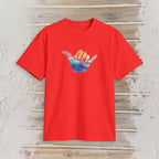 A bright red short-sleeve t-shirt with a colorful graphic of a hand making a shaka sign over a sunset and ocean scene, displayed flat on a rustic light wood background.