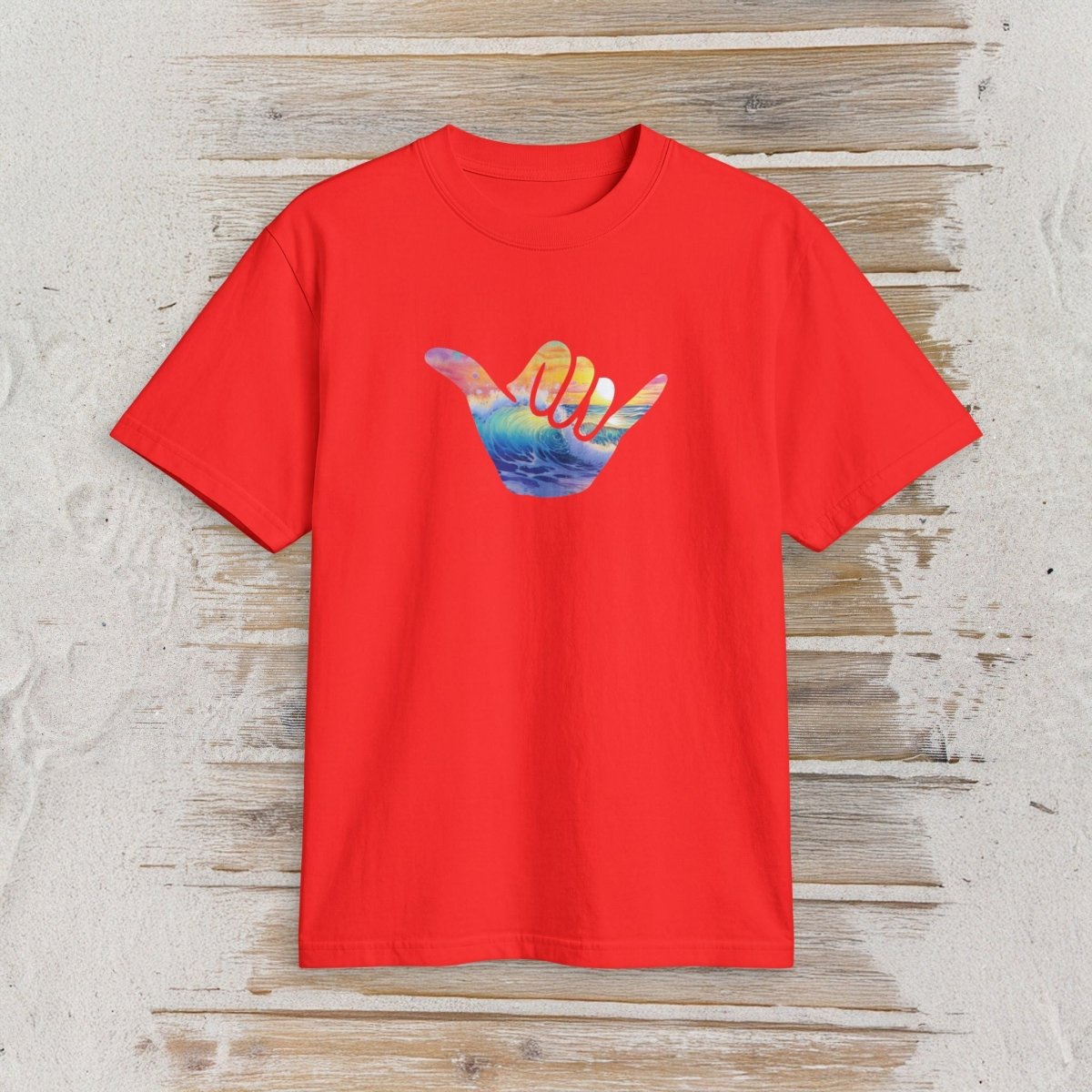 A bright red short-sleeve t-shirt with a colorful graphic of a hand making a shaka sign over a sunset and ocean scene, displayed flat on a rustic light wood background.