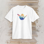 A white short-sleeve crewneck t-shirt featuring a colorful graphic of a hand in a shaka sign gesture, with the palm side showing a watercolor-style rainbow gradient. The t-shirt is displayed flat against a rustic, light-colored wooden plank background.
