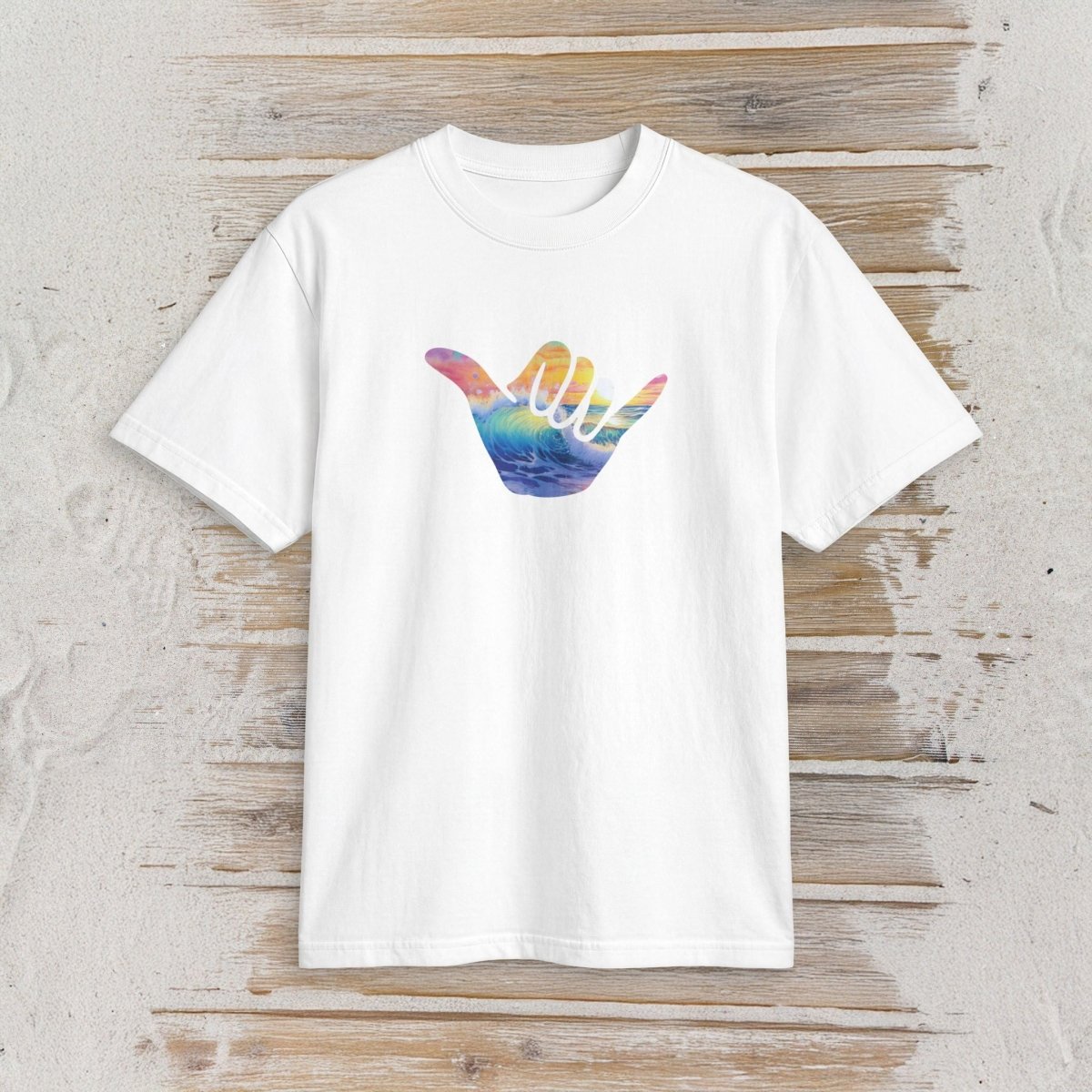 A white short-sleeve crewneck t-shirt featuring a colorful graphic of a hand in a shaka sign gesture, with the palm side showing a watercolor-style rainbow gradient. The t-shirt is displayed flat against a rustic, light-colored wooden plank background.