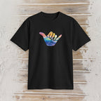 A black short-sleeve crewneck t-shirt featuring a colorful Hawaiian 'shaka' sign graphic on the chest, displayed flat against a rustic light wood plank background.