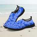 A pair of slip-on water shoes with a blue shark pattern and black soles, displayed on a sandy beach with the ocean in the background.