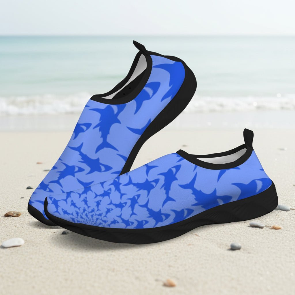 A pair of slip-on water shoes with a blue shark pattern and black soles, displayed on a sandy beach with the ocean in the background.