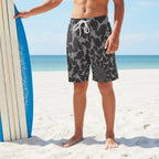 Teen surfer dude wearing black swim shorts with gray sharks on a beach with a surfboard.