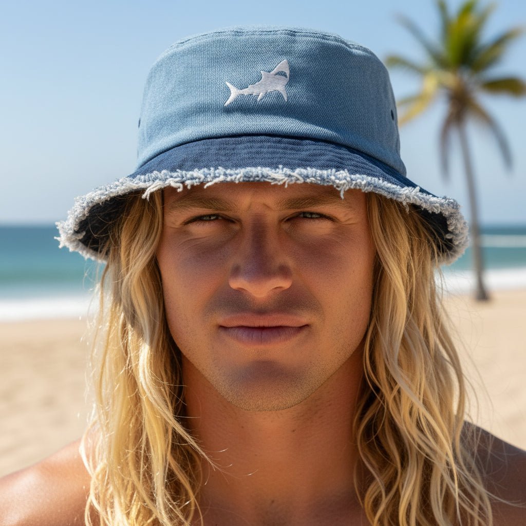A man with long blonde hair wearing a light blue bucket hat with a white shark embroidery, standing on a sandy beach with the ocean and a palm tree in the background.