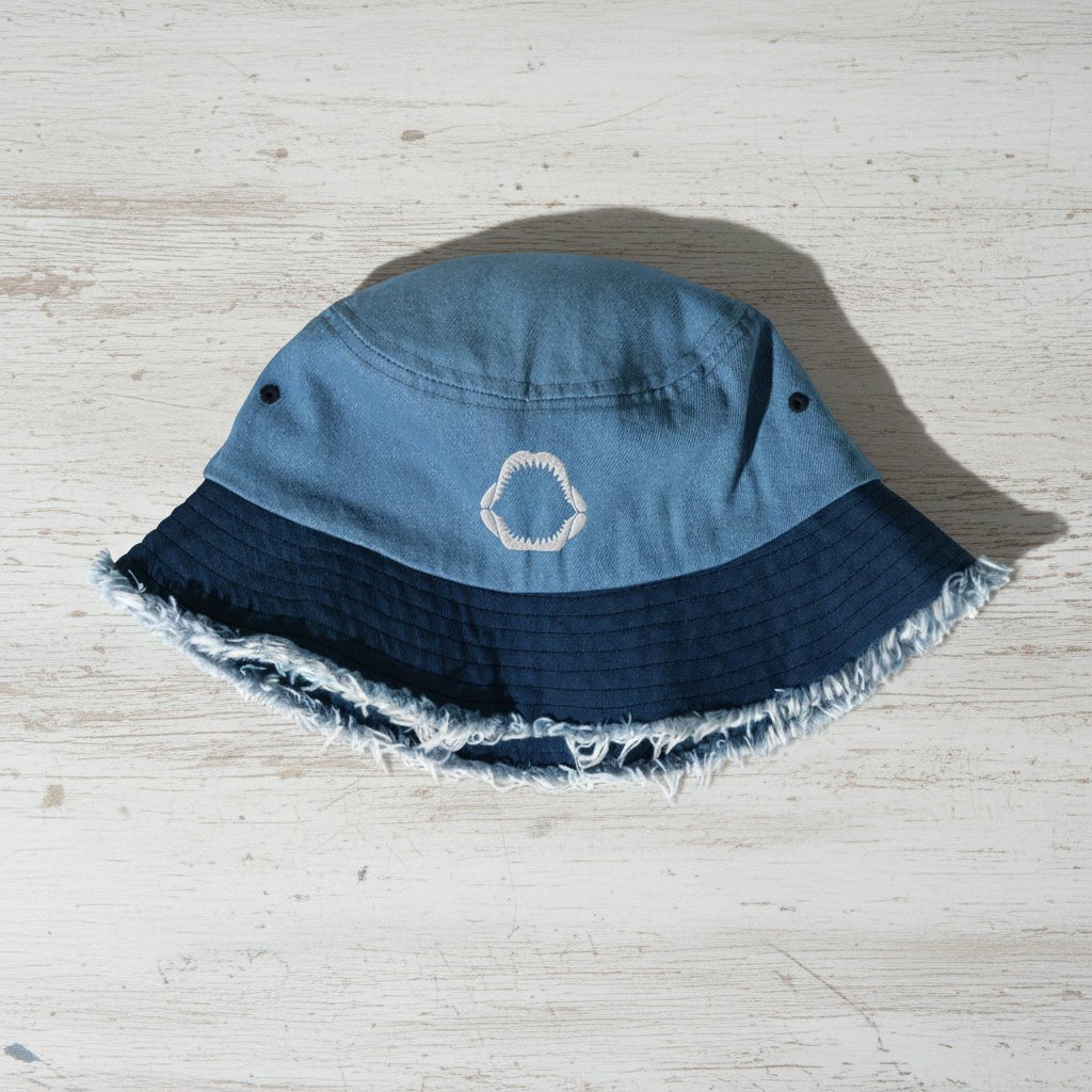 Blue bucket hat with frayed edge and embroidered shark jaw a nautical wood surface