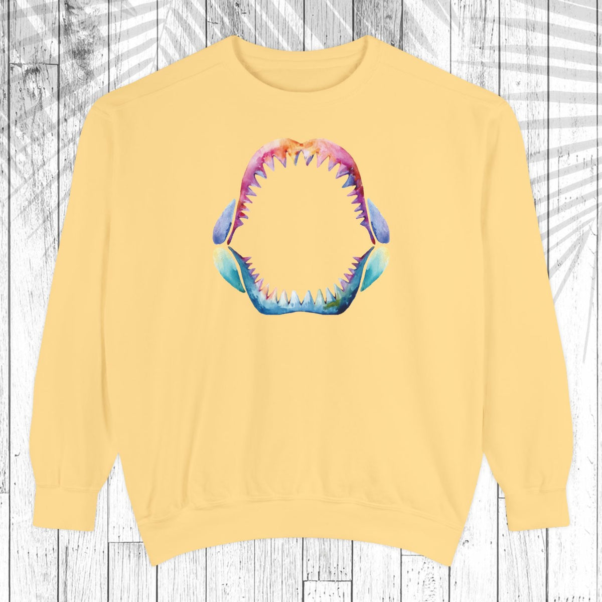 A pale yellow crewneck sweatshirt featuring a colorful graphic of an open shark mouth with sharp teeth on the chest, displayed against a white wood plank background.